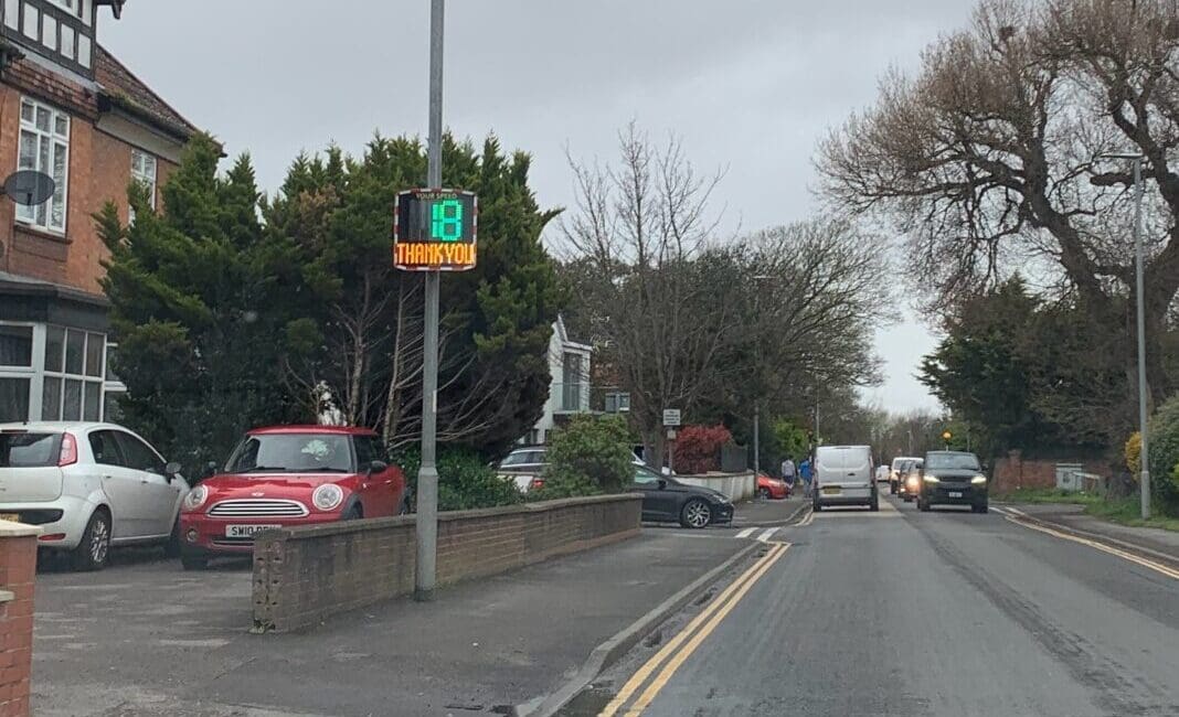 A Speed Indicator Device (SID) displaying "18" and "THANK YOU!" installed on a residential street in Burnham-On-Sea, UK, with cars parked on the side and driving on the road.