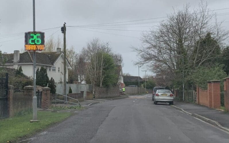 A Speed Indicator Device (SID) displaying "26" and "THANK YOU!" installed on a residential street in Burnham-On-Sea, UK, with houses and a parked car visible in the background.